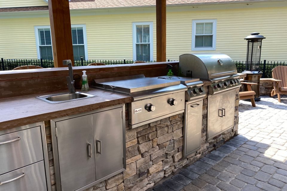 Outdoor kitchen counter in stone with a gas grill, griddle and sink beside a paver patio