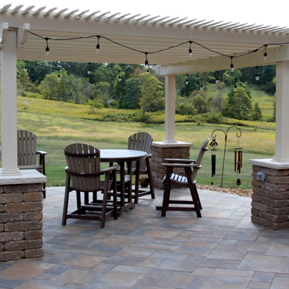 White pergola on stone-clad columns over a paver patio, with string lights above the table