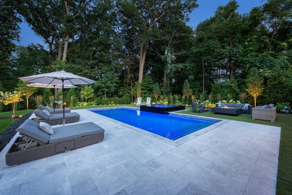 Pale stone pool deck around a rectangular pool, with loungers and a lit garden beyond at dusk