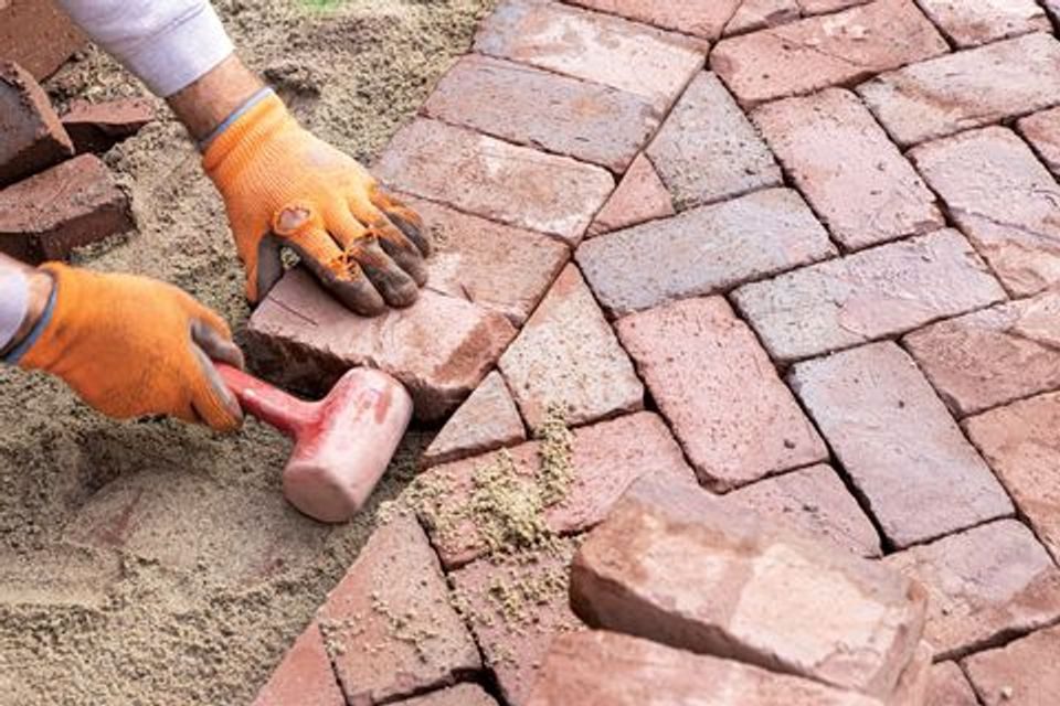 Gloved hands bedding a clay brick paver into sand with a rubber mallet