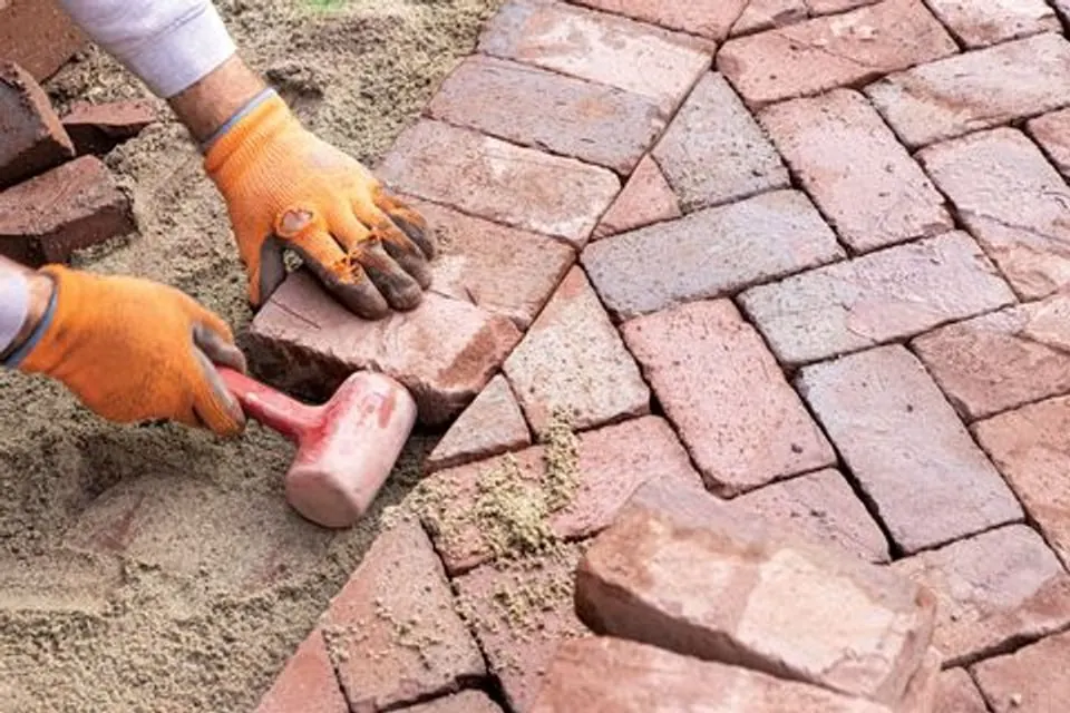 Gloved hands bedding a clay brick paver into sand with a rubber mallet