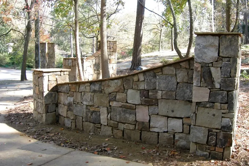 Dry-stacked stone wall with square capped columns running along a wooded drive