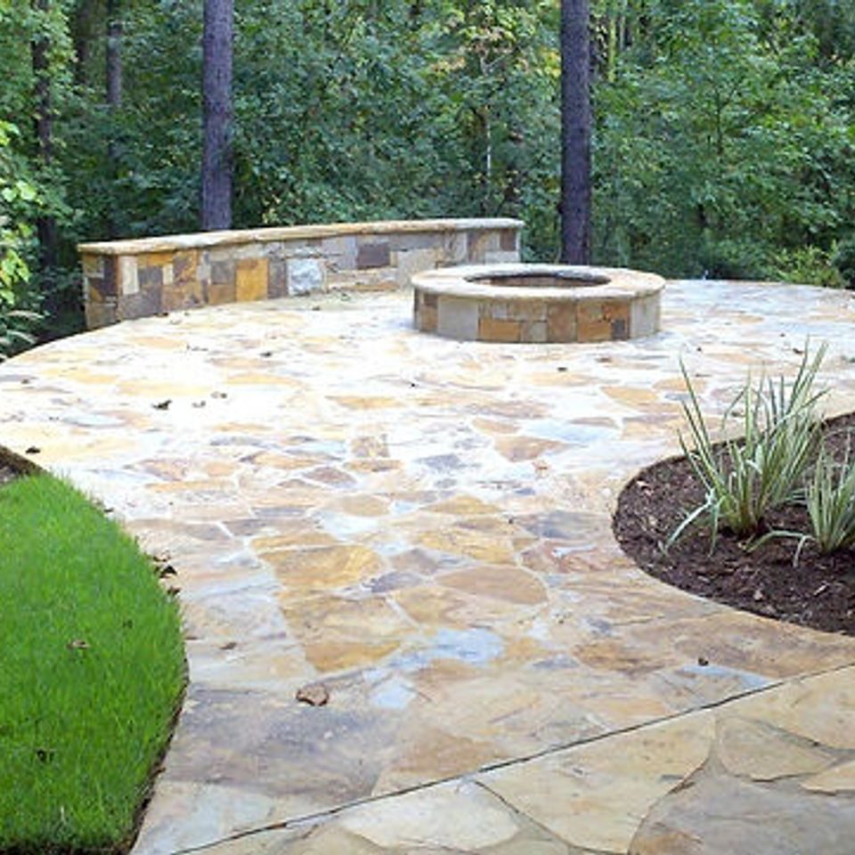 Tan flagstone patio curving around a stone fire pit, with a seat wall along the woodland edge