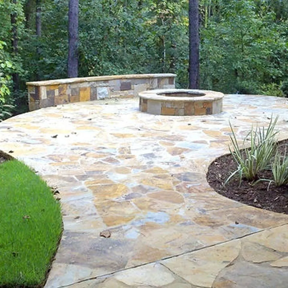Tan flagstone patio curving around a stone fire pit, with a seat wall along the woodland edge