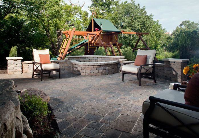 Block fire pit and seat wall on a paver patio, with a children's play set beyond