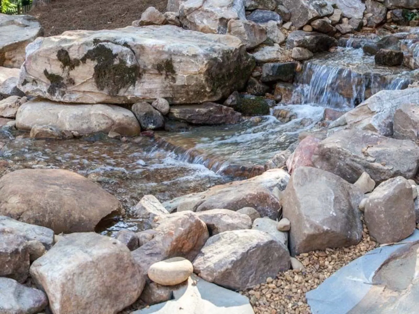 Rock-lined stream and low waterfall running between set boulders and washed river gravel