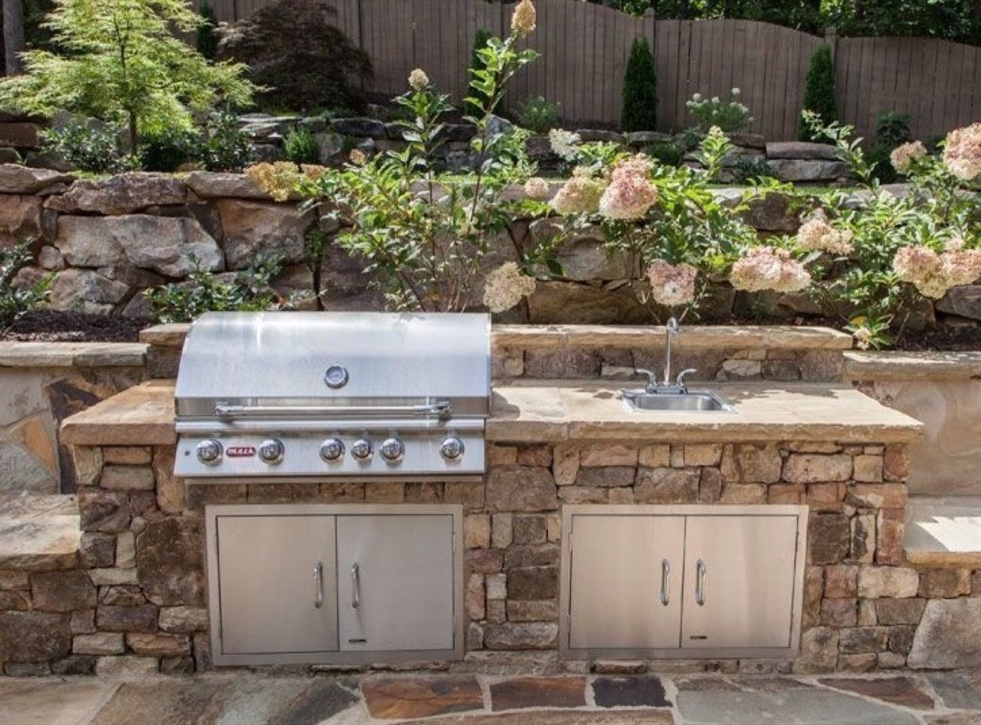 Natural stone outdoor kitchen with a built-in grill and sink below a boulder wall planted with hydrangeas