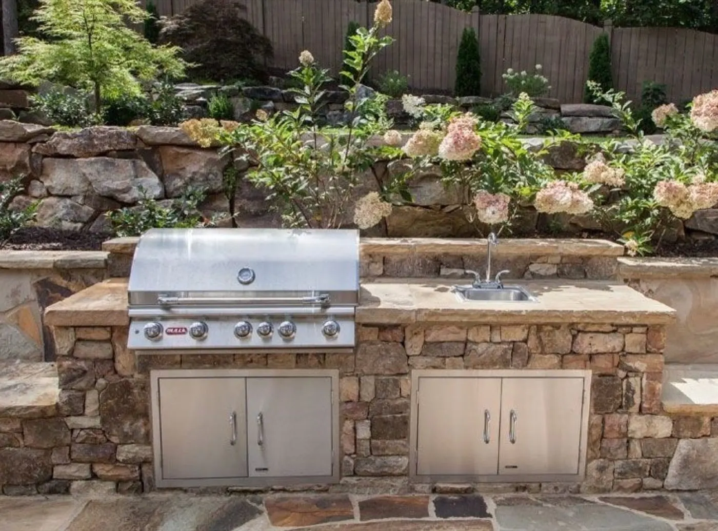 Natural stone outdoor kitchen with a built-in grill and sink below a boulder wall planted with hydrangeas