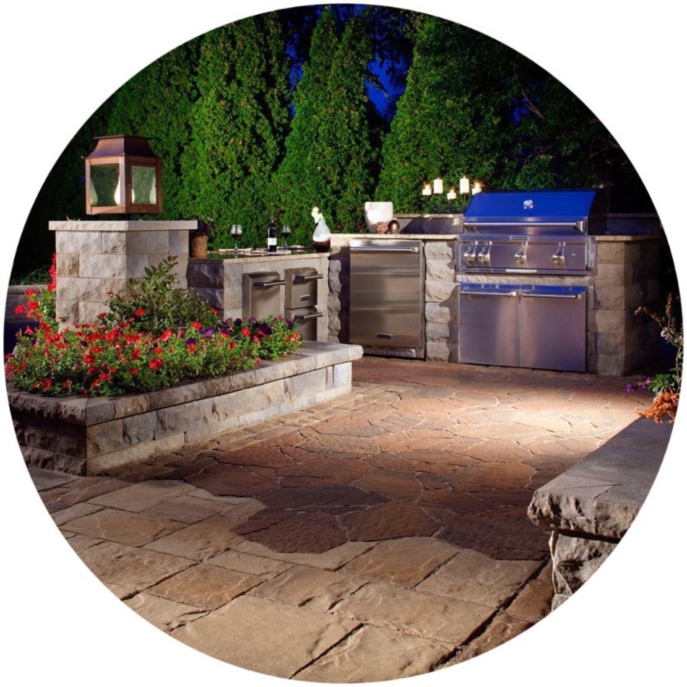 Outdoor kitchen with a built-in grill and fridge lit at night above a flagstone-look paver patio