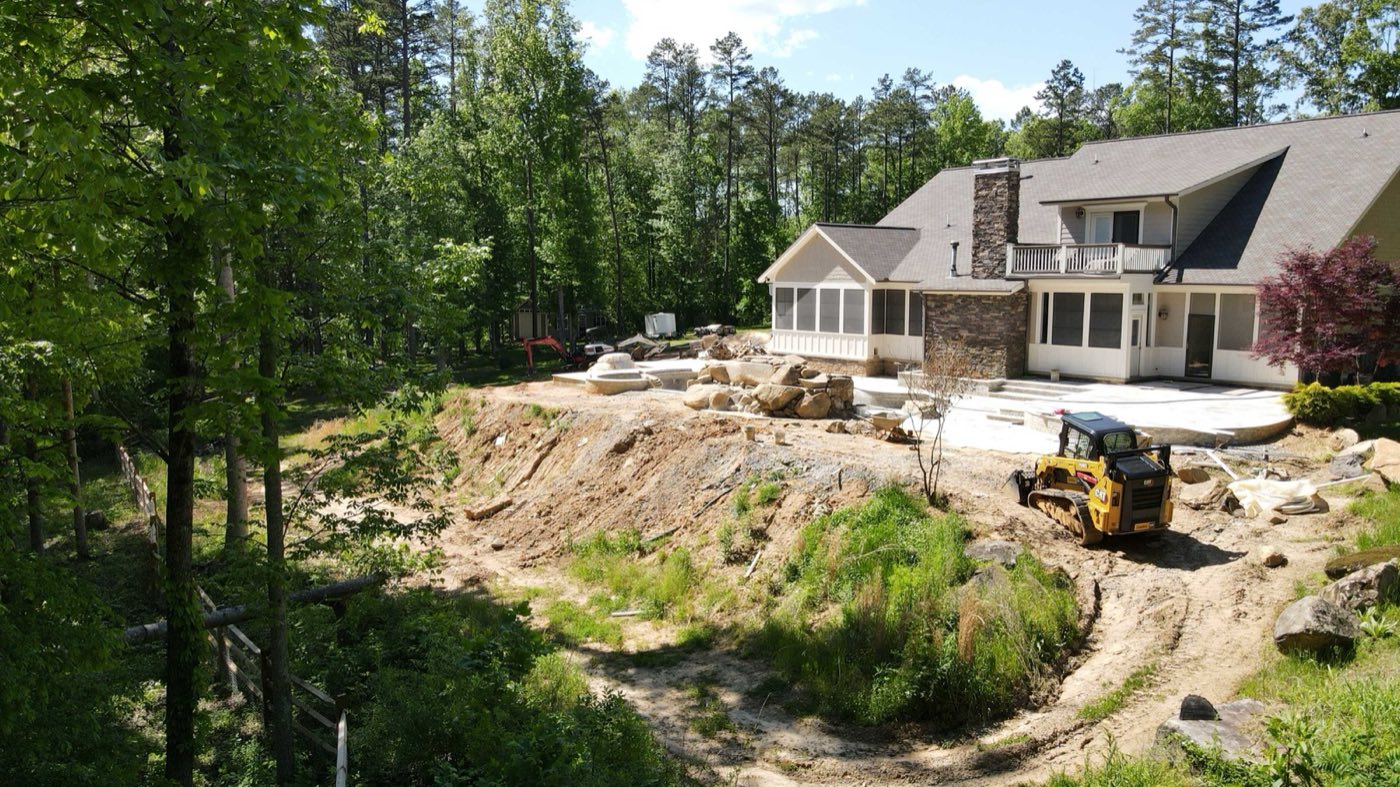 Excavated hillside behind a Concord home with boulders staged and a skid steer working the grade
