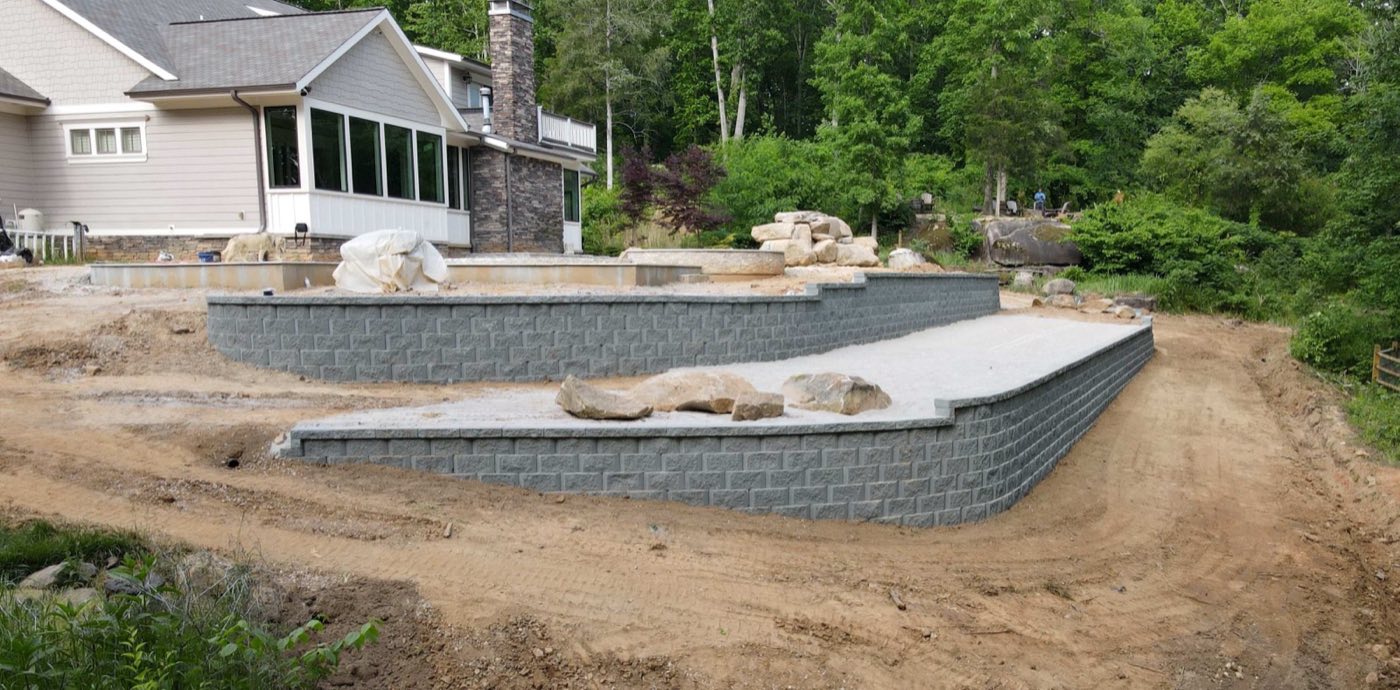 Two tiers of grey segmental block retaining wall curving around a new concrete pad in Concord