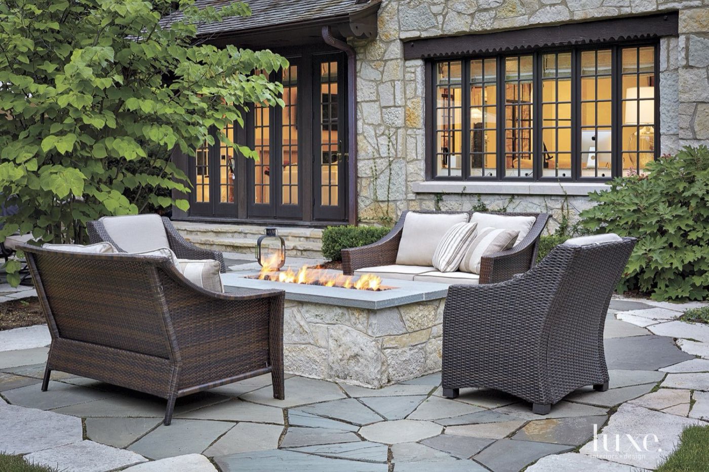 Square gas fire table with a stone base on an irregular flagstone patio beside a stone house