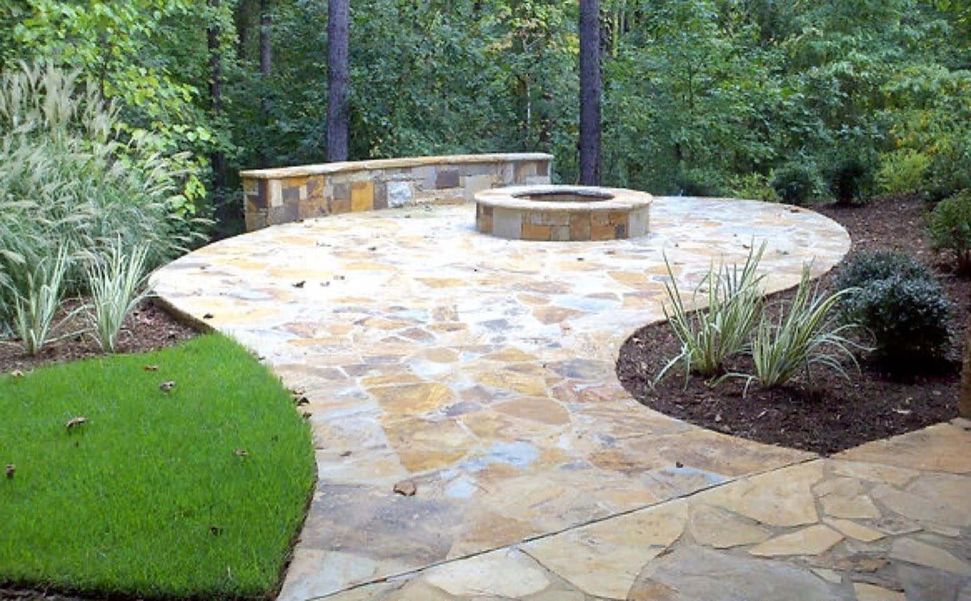 Curved tan flagstone patio with a round fire pit and a low stone seat wall at the woodland edge