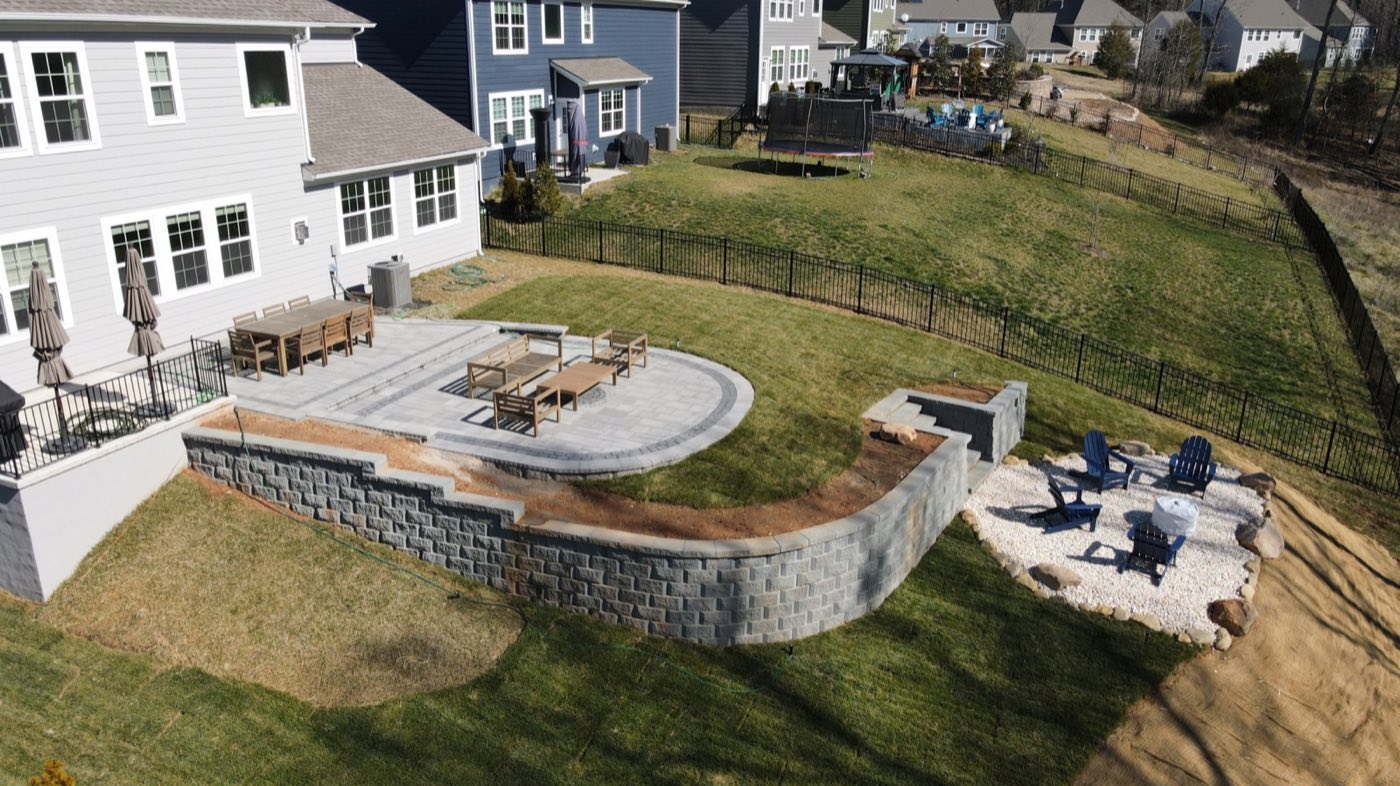 Aerial of a raised patio held by a block retaining wall, with steps down to a gravel fire pit circle