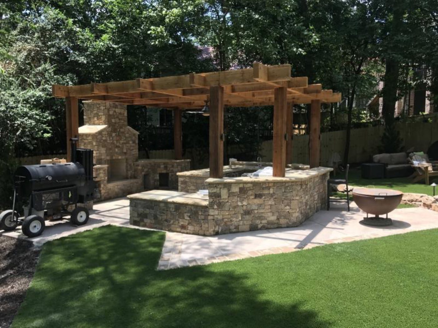 Stone bar and kitchen island under a timber pergola, with an outdoor fireplace and copper fire bowl