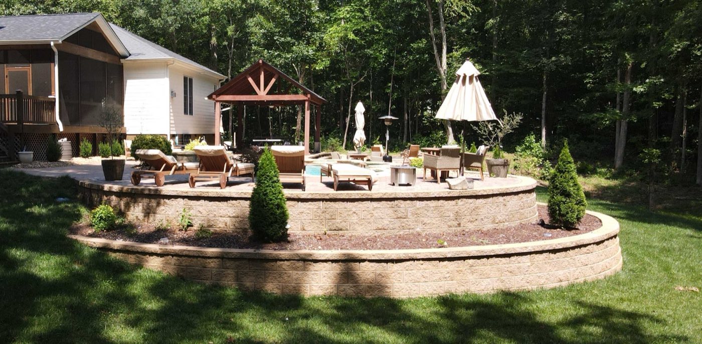 Raised patio held by a curved block wall, with a hot tub and pergola above the lawn