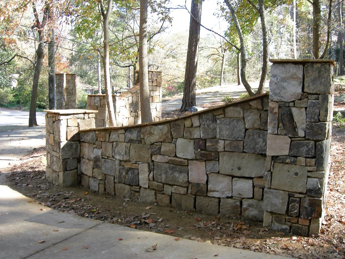 Dry-stacked stone entrance wall stepping between square stone columns along a drive